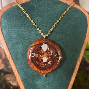 Op de afbeelding: Een ketting met een ronde hanger van gepolijst houtschijfje aan een goudkleurige ketting. De hanger is ingesloten in heldere hars en versierd met twee kleine ovale maansteen cabochons en vier goudkleurige maansikkel bedels.