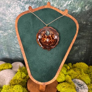 May include: A circular pendant made from a dark brown pine cone slice, featuring a central oval gemstone, two smaller round stones, and a small silver star charm. The pendant is suspended on a thin gold-toned chain and displayed on a green stand.