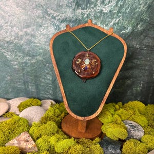 Op de afbeelding: Een ketting met een gouden ketting en een houten hanger. De ronde hanger heeft een donkere houtnerf en ingebedde edelstenen. De ketting wordt getoond op een houten standaard met een groene fluwelen achterkant, tegen een achtergrond van groen mos en stenen.