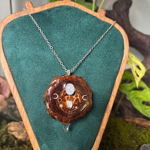 Op de afbeelding: Een zilveren ketting met een ronde hanger van gepolijst hout. De hanger is ingelegd met twee ovale maanstenen, kleine metalen cirkelvormige accenten en twee maansikkel-symbolen, allemaal afgewerkt met een glanzende laag van hars.