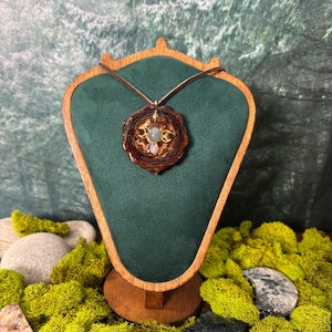 May include: A handcrafted pendant necklace with a brown leather cord. The pendant features a circular, wooden design with gold accents and a central light blue gemstone. The necklace is displayed on a green velvet stand, surrounded by green moss.