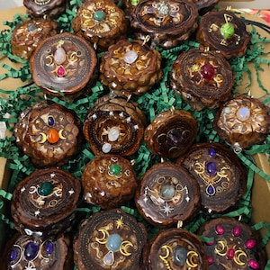 Peut inclure: Une collection de pendentifs artisanaux en pommes de pin, chacun orné d'une variété de pierres précieuses et de motifs célestes. Les pendentifs présentent des accents dorés et sont présentés dans une boîte en carton avec du papier vert.