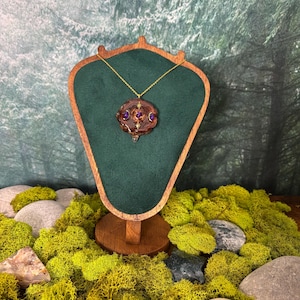Puede incluir: Un collar con colgante artesanal con una cadena dorada. El colgante tiene una forma orgánica marrón oscuro con piedras preciosas moradas incrustadas. El collar se exhibe en un expositor de joyas de madera con respaldo de terciopelo verde.