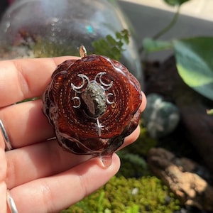 May include: A circular, dark brown, polished pine cone slice pendant featuring a central, rough, metallic-toned stone. The stone is surrounded by a silver-colored metal inlay depicting a triple moon phase symbol and a small star at the bottom.
