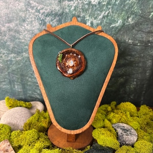 Op de afbeelding: Een ronde houten hanger met een centrale iriserende maansteen cabochon, omringd door kleine zilveren steraccenten en een klein groen blaadje als bedel, hangend aan een dun bruin koord op een donkergroene fluwelen displaystandaard.