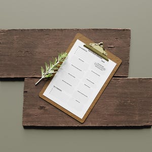 Op de afbeelding: Een klembord met een afgedrukte lijst met dagelijkse intenties, rustend op twee verweerde houten planken. Een takje groen is bij het klembord geplaatst. Het klembord heeft een goudkleurige clip aan de bovenkant.