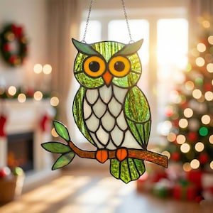 Puede incluir: Decoración de búho de vidriera con tonos verdes, blancos y naranjas. El búho está posado en una rama marrón con hojas verdes. El búho tiene grandes ojos amarillos y cuelga de una cadena. El fondo incluye un árbol de Navidad y una chimenea.