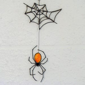 May include: A stained glass hanging ornament featuring a black spider with a bright orange abdomen suspended by a thin wire from a black-framed spider web. The web is constructed with clear, textured glass panels held together by dark metal solder lines.