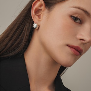 May include: A close-up of a person wearing a black blazer and silver ball stud earrings. The earrings are round with a textured surface. A delicate silver necklace is also visible.
