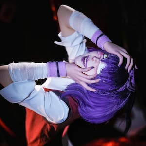 May include: A person with purple hair and makeup, wearing a white shirt with red accents and bandages on the arm. The person is wearing purple wristbands and has purple nail polish. The background is dark.