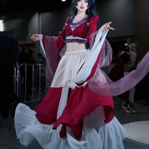 Op de afbeelding: Een vrouw in een cosplay-kostuum met een rode en witte jurk met zwierige mouwen en een gelaagde rok. De outfit bevat een rode crop top, een witte rok en een paarse accent. De vrouw heeft donkergroen haar en een bloem in haar haar.