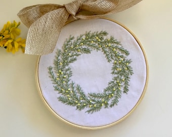 Könnte beinhalten: Handgestickter Blumenkranz in einem Holzreifen. Das Design zeigt grüne Blätter und gelbe Akzente auf weißem Stoff. Darüber befindet sich eine Schleife aus Jute. Gelbe Blumen im Hintergrund. Durchmesser ca. 20 cm.