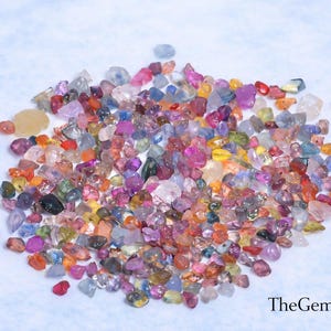 May include: A collection of multi-coloured gemstones in various shapes and sizes. The gems display a range of colours, including pink, purple, orange, yellow, and blue. The gemstones are scattered on a light blue surface.