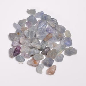 May include: A collection of raw, unpolished gemstones in various shades of light blue, lavender, and pale peach. These small, irregular mineral fragments feature translucent to opaque surfaces with natural facets and smooth, rounded edges throughout.