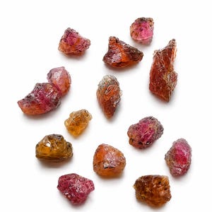 May include: A collection of twelve raw, unpolished gemstones in varying shades of deep red, orange, and amber. Each stone features a natural, irregular, and faceted texture with translucent qualities, scattered across a plain white surface.