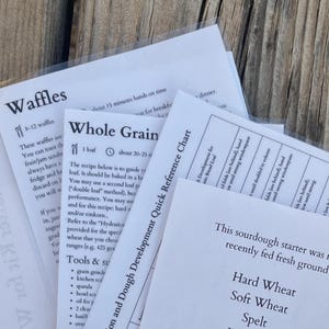 May include: A collection of white recipe cards with black text, including titles such as "Waffles" and "Whole Grain." The cards are in clear plastic sleeves, set against a wooden background.