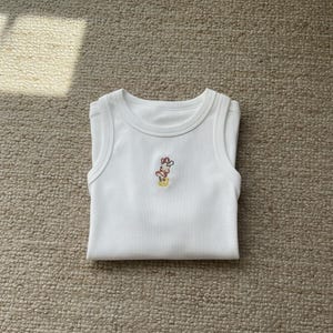 May include: White baby tank top with a small embroidered design of a cartoon character. The tank top is folded and laying on a neutral-colored carpet. The embroidered design features red, yellow, and brown colors.