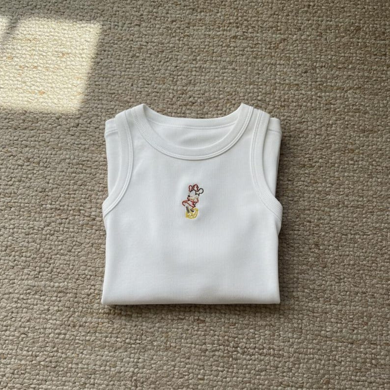 May include: White baby tank top with a small embroidered design of a cartoon character. The tank top is folded and laying on a neutral-colored carpet. The embroidered design features red, yellow, and brown colors.