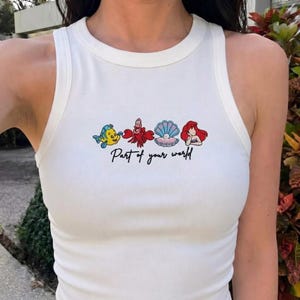 May include: White tank top with a crew neck featuring embroidered cartoon characters and the text "Part of your world" in black script. The design includes a yellow fish, a red crab, a seashell, and a red-haired mermaid.
