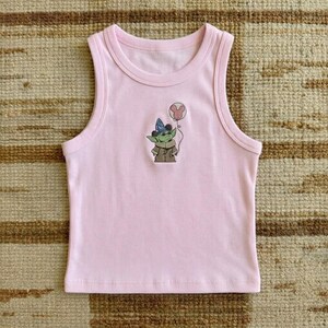 Pu&ograve; includere: Una canotta rosa chiaro con scollo tondo e una grafica a cartoni animati di un personaggio verde con un cappello da mago e un palloncino di Topolino. La canotta &egrave; realizzata in un materiale morbido e a coste.