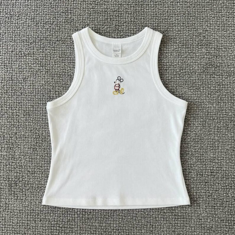 May include: White tank top with a small embroidered Mickey Mouse design. The sleeveless top has a round neckline and a fitted silhouette. The Mickey Mouse design features black, red, yellow, and white colors.