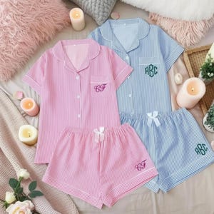 Puede incluir: Se muestran dos conjuntos de pijama a rayas, uno rosa y otro azul. Cada conjunto incluye una camisa abotonada de manga corta y pantalones cortos a juego. El conjunto rosa tiene "CF" bordado en el bolsillo, mientras que el azul tiene "ABC". Lazos blancos adornan los pantalones cortos.