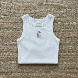 May include: White ribbed tank top with a small embroidered Mickey Mouse design in the center. The sleeveless top has a round neckline and is folded neatly. The embroidery features yellow, black, and pink colors.