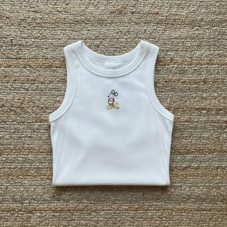 May include: White ribbed tank top with a small embroidered Mickey Mouse design in the center. The sleeveless top has a round neckline and is folded neatly. The embroidery features yellow, black, and pink colors.