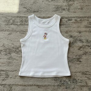 May include: White ribbed tank top with a small embroidered design near the neckline. The design features a cartoon character with a red bow. The top has a scoop neck and a fitted silhouette.