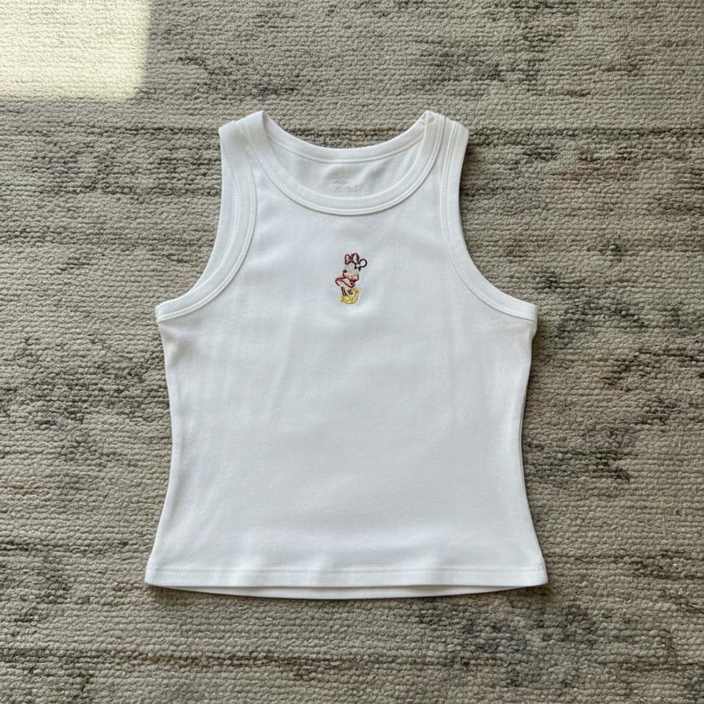May include: White ribbed tank top with a small embroidered design near the neckline. The design features a cartoon character with a red bow. The top has a scoop neck and a fitted silhouette.
