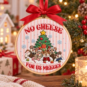 Op de afbeelding: Een rond borduurringornament met een lichtroze en wit gestreepte stoffen achtergrond. Het borduurwerk toont vijf muizen verzameld rond een versierde kerstboom met cadeautjes, sneeuwvlokken en de tekst NO CHEESE FOR US MEEGES.