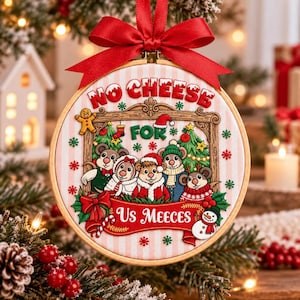 Könnte beinhalten: Ein rundes Stickrahmen-Ornament aus Holz mit einem hellrosa und weiß gestreiften Stoffhintergrund. Die Stickerei zeigt fünf Mäuse in Winterkleidung, einen Lebkuchenmann und einen Schneemann mit dem Text NO CHEESE FOR US MEECES.