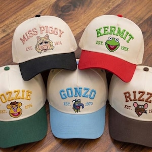 May include: Five baseball caps with off-white crowns and colored brims. Each cap features embroidered character names and faces: Miss Piggy with a black brim, Kermit with a red brim, Fozzie with a green brim, Gonzo with a light blue brim, and Rizzo with a dark brown brim, all including establishment years.
