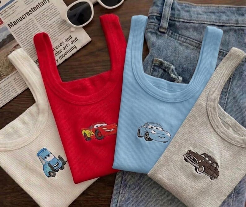 K&ouml;nnte beinhalten: Vier gerippte Tanktops in Wei&szlig;, Rot, Hellblau und Grau, jedes mit einem Cartoon-Auto-Design. Die Oberteile werden auf einem Jeanshintergrund mit einer Zeitung und einer Sonnenbrille pr&auml;sentiert.
