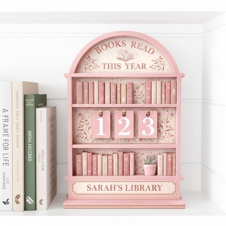 Puede incluir: Una estanter&iacute;a de madera rosa con el texto "BOOKS READ THIS YEAR" y "SARAH'S LIBRARY". La estanter&iacute;a tiene tres estantes llenos de libros en miniatura y tres etiquetas numeradas. Una peque&ntilde;a planta en maceta se encuentra en el estante inferior. Libros a la izquierda.