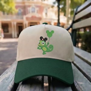 May include: A baseball cap with a green brim and a tan crown. The cap features an embroidered green chameleon holding a green Mickey Mouse balloon. The chameleon wears a black bow and a black Mickey Mouse ear hat.