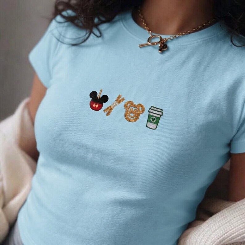 May include: Light blue t-shirt with embroidered icons of a Mickey Mouse head, pretzel sticks, a Mickey Mouse pretzel, and a coffee cup. The shirt is short-sleeved and has a crew neck. A gold necklace is visible.