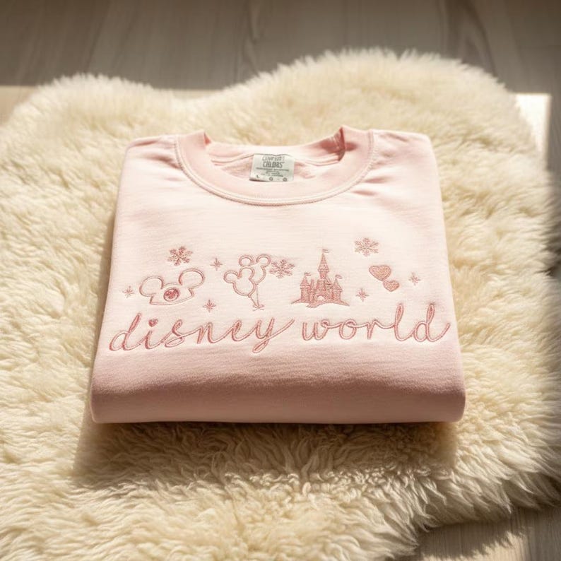 Peut inclure: Un sweat-shirt rose clair avec l'inscription brod&eacute;e "disney world" en cursive. Au-dessus du texte, des ic&ocirc;nes brod&eacute;es sur le th&egrave;me de Disney, dont Mickey Mouse et un ch&acirc;teau. Le sweat-shirt est pli&eacute; sur une surface moelleuse de couleur cr&egrave;me.