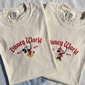 May include: Two off-white t-shirts with the embroidered text "Disney World" in red script, along with the year "1971". One shirt features Mickey Mouse, the other Minnie Mouse, both holding paintbrushes. The shirts are laid on a white surface.