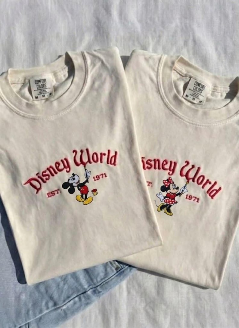 May include: Two off-white t-shirts with the embroidered text "Disney World" in red script, along with the year "1971". One shirt features Mickey Mouse, the other Minnie Mouse, both holding paintbrushes. The shirts are laid on a white surface.