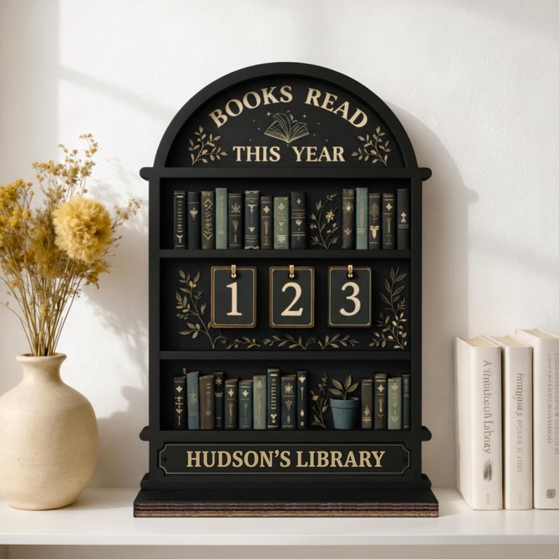 Puede incluir: Estanter&iacute;a de madera negra con el texto "BOOKS READ THIS YEAR" y "HUDSON'S LIBRARY". La estanter&iacute;a tiene tres estantes llenos de libros en miniatura y un contador para llevar un registro de los libros le&iacute;dos.