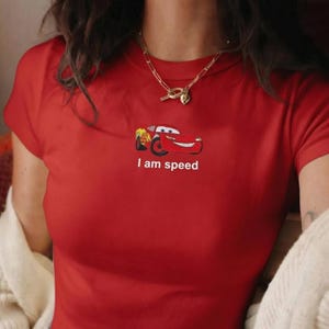 May include: Red t-shirt featuring a cartoon car graphic with the text "I am speed" in white. The short-sleeved shirt has a crew neck and is made of a soft material. A gold necklace is visible.