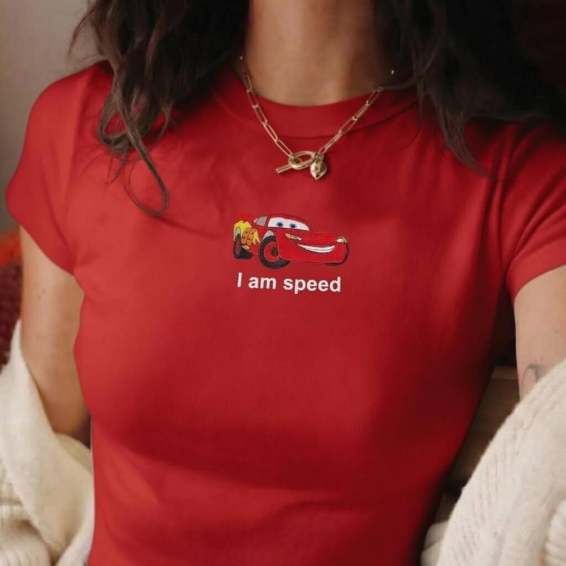 May include: Red t-shirt featuring a cartoon car graphic with the text "I am speed" in white. The short-sleeved shirt has a crew neck and is made of a soft material. A gold necklace is visible.