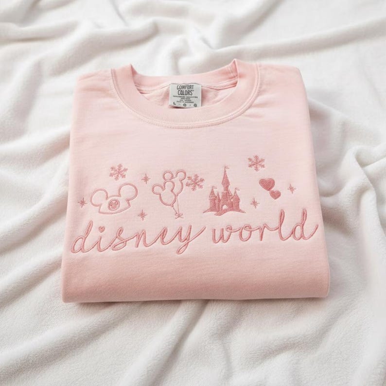 Peut inclure: Un sweat-shirt rose clair avec des broderies Disney World. Le motif comprend Mickey Mouse, des ballons, un ch&acirc;teau, des flocons de neige et des c&oelig;urs. Les mots "disney world" sont brod&eacute;s en cursive.
