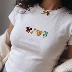 May include: White t-shirt with embroidered designs of a Mickey Mouse-shaped candy apple, pretzel sticks, a Mickey Mouse-shaped pretzel, and a coffee cup. The shirt has a crew neck and short sleeves.
