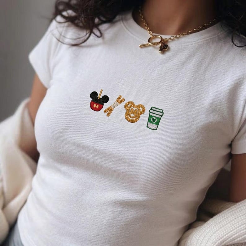 May include: White t-shirt with embroidered designs of a Mickey Mouse-shaped candy apple, pretzel sticks, a Mickey Mouse-shaped pretzel, and a coffee cup. The shirt has a crew neck and short sleeves.
