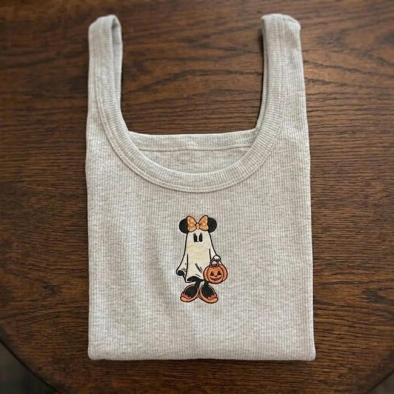 May include: A light gray ribbed tank top featuring a central embroidered graphic of a ghost character wearing a bow and holding a small orange pumpkin. The ghost has black ears, orange accents on the bow and shoes, and a carved jack-o'-lantern face.
