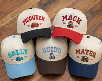 May include: Five baseball caps with embroidered designs. The caps feature the names "McQueen," "Mack," "Sally," "Guido," and "Mater," along with car illustrations and the years 1977, 1949, 2002, 2006, and 1951. Each cap has a different color scheme.