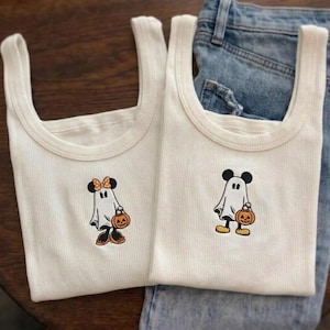 May include: Two white ribbed tank tops featuring embroidered ghost characters. One ghost wears a polka-dot bow and holds a pumpkin, while the other ghost wears round ears and holds a pumpkin. The fabric is a textured ribbed knit material.