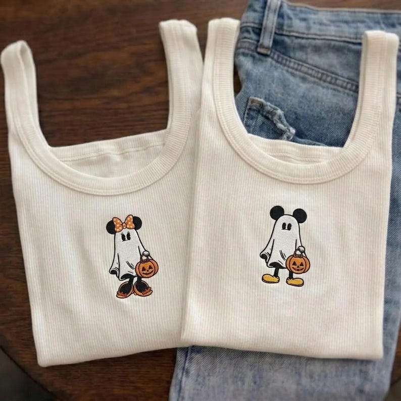 May include: Two white ribbed tank tops featuring embroidered ghost characters. One ghost wears a polka-dot bow and holds a pumpkin, while the other ghost wears round ears and holds a pumpkin. The fabric is a textured ribbed knit material.